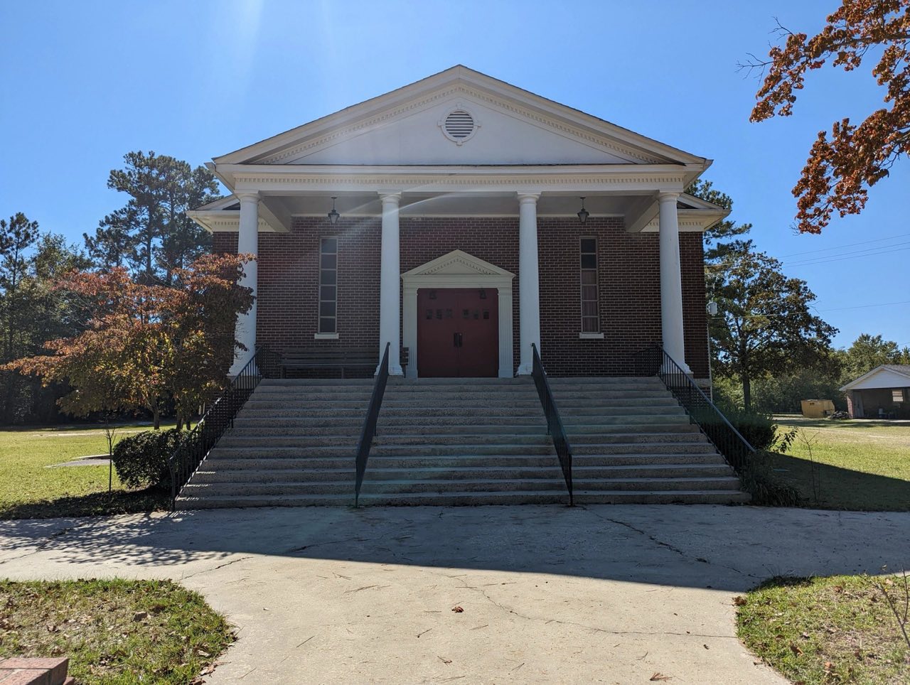 The front of Hillcrest Church: red brick, four white columns and red double doors at the top of wide steps, under a blue sky